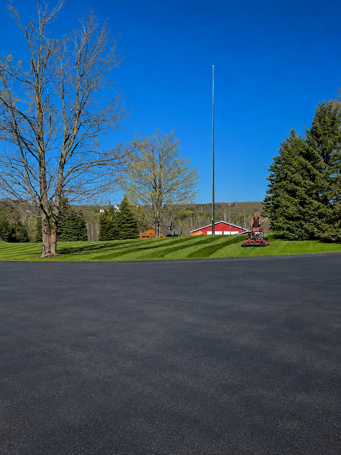 Paved lot beside grassy park with trees, a flagpole, and a red-and-white building under a clear blue sky
