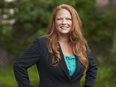 Smiling woman in a black blazer and teal blouse standing outdoors with a blurred green background