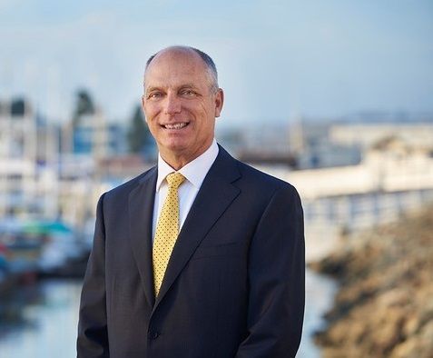 Man in a dark suit and yellow tie standing outdoors with a blurred cityscape behind him.
