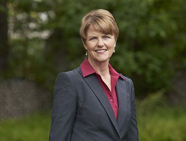 Smiling woman in a dark gray blazer and red shirt standing outdoors against a blurred green background