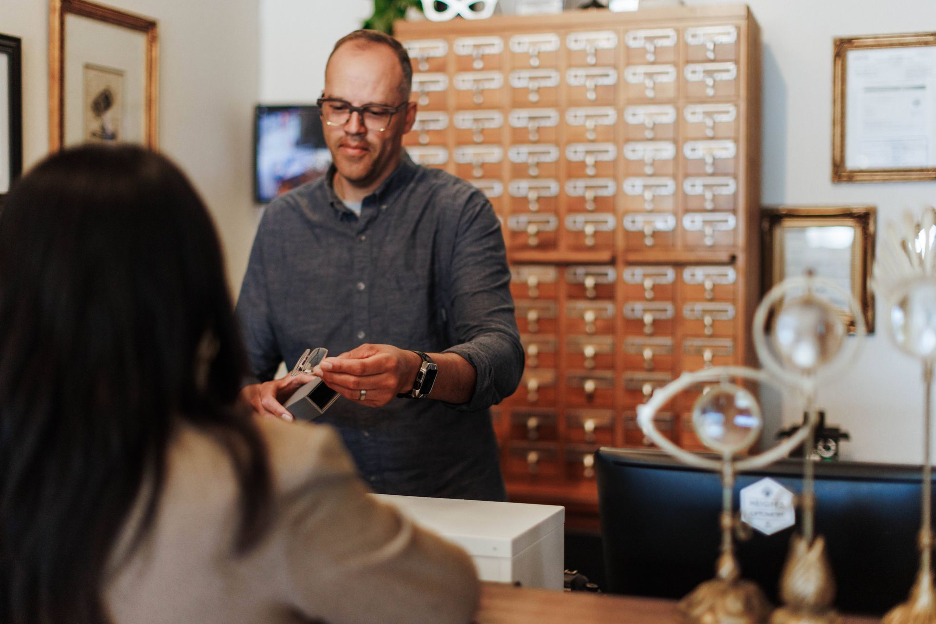 A person wearing glasses and a dark shirt hands a small item to a customer at a counter in front of a wooden filing cabinet.