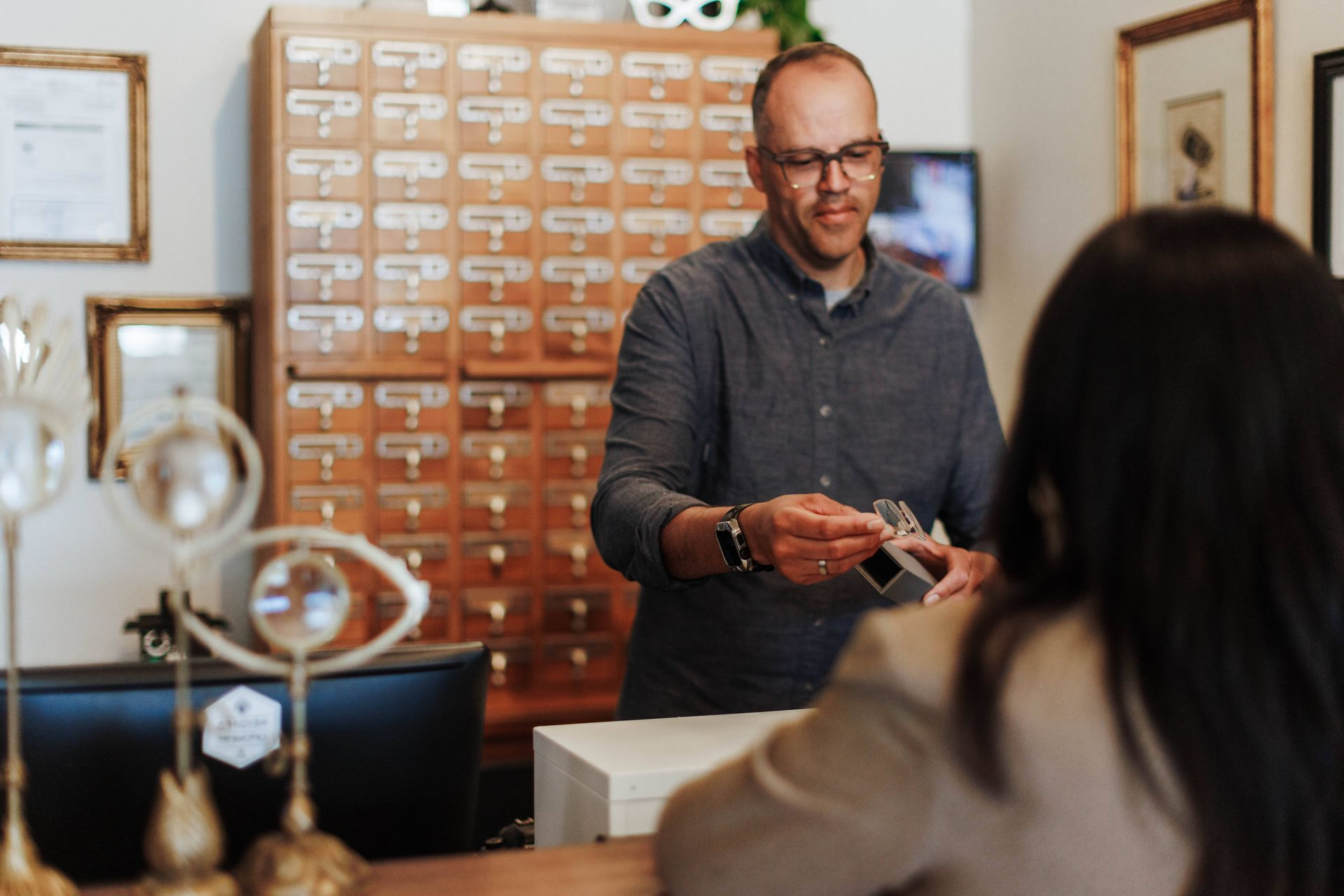 A person in a gray shirt stands behind a counter, handing a small item to a customer in an eye care office.