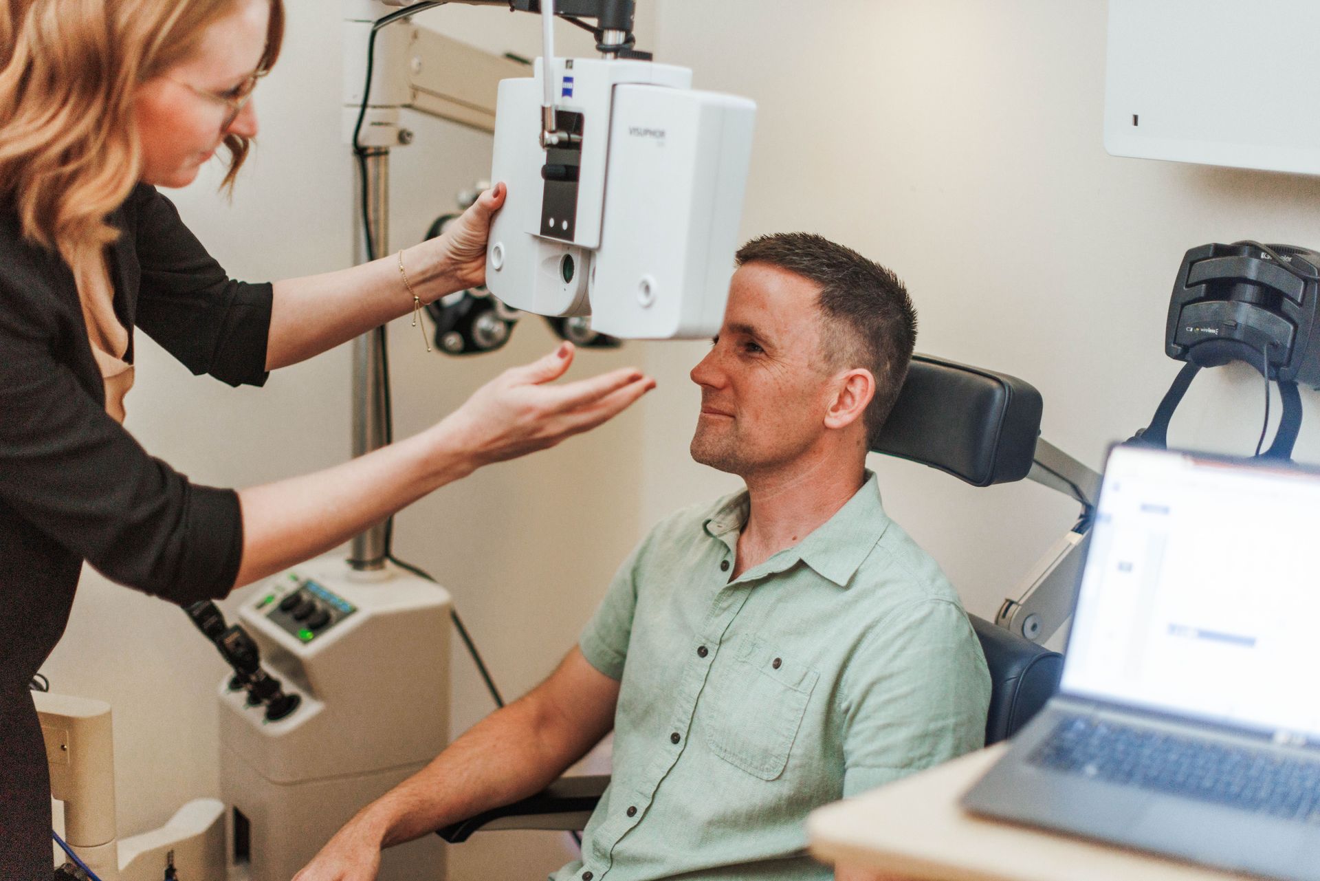 An optometrist adjusts a phoropter in front of a patient seated in an exam chair for a vision test.