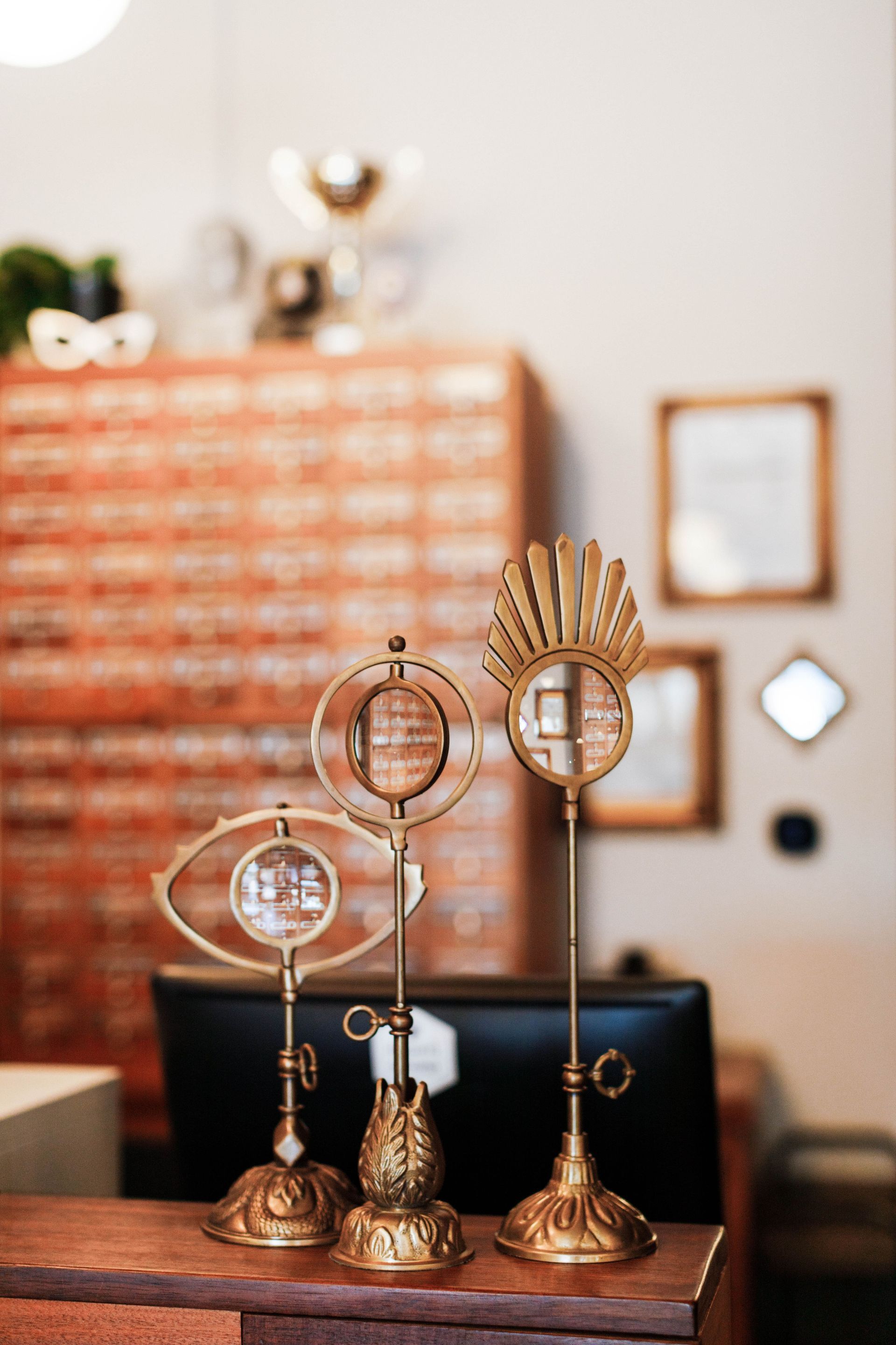Three ornamental gold-toned magnifying glasses on stands sit on a wooden counter in front of a grid of small drawers.