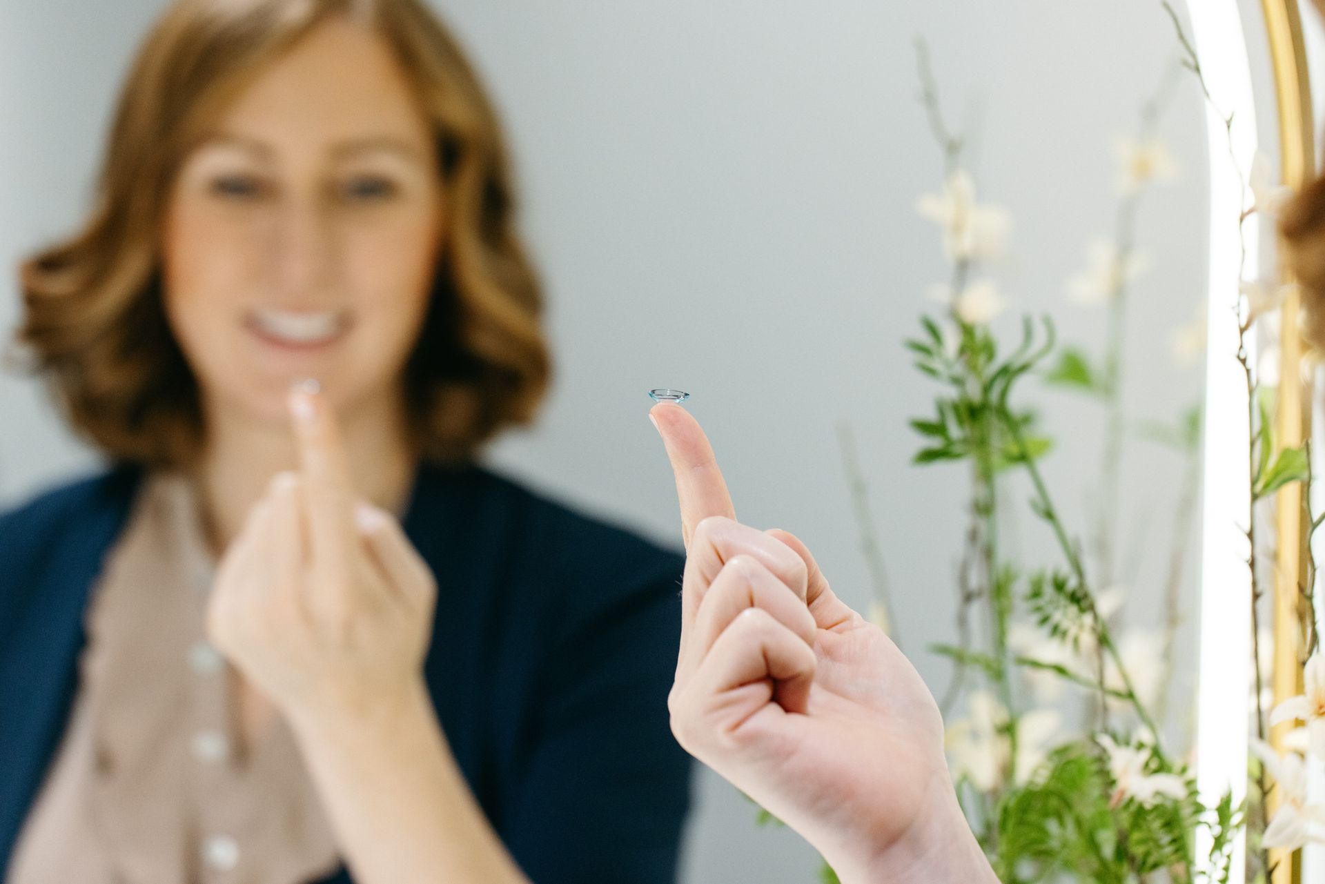 A person holds a small contact lens on their fingertip in front of a mirror, with their reflection visible.