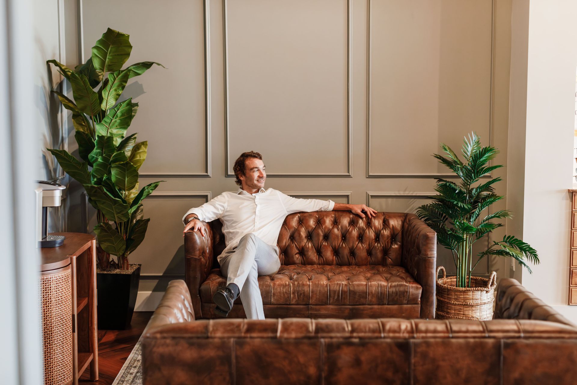 A person sits comfortably on a brown tufted leather sofa in a room with paneled walls, tall indoor plants, and wooden decor.