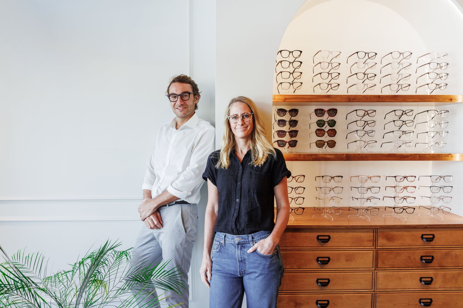 Two people standing beside a wooden display cabinet filled with various eyeglasses in a brightly lit, minimalist shop.