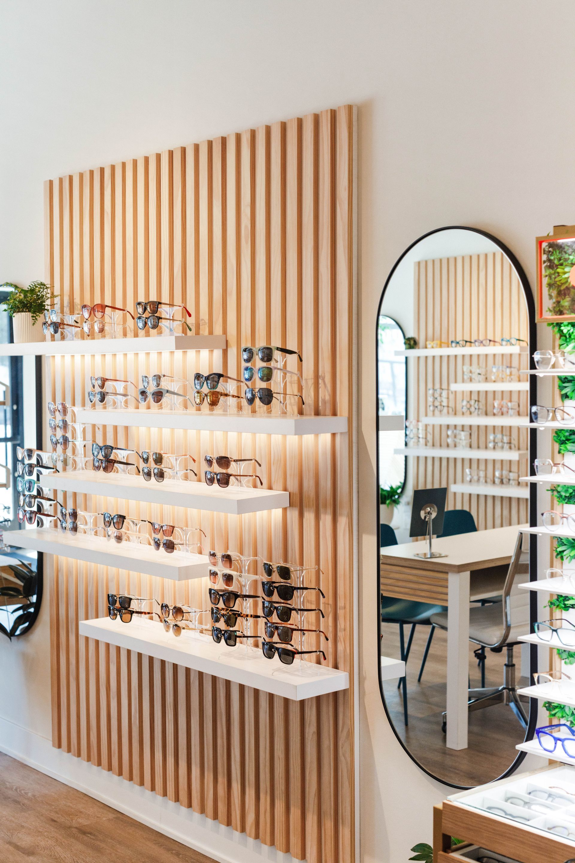 A bright optical store display with eyewear on illuminated wooden shelves and a tall oval mirror on a slatted wall.