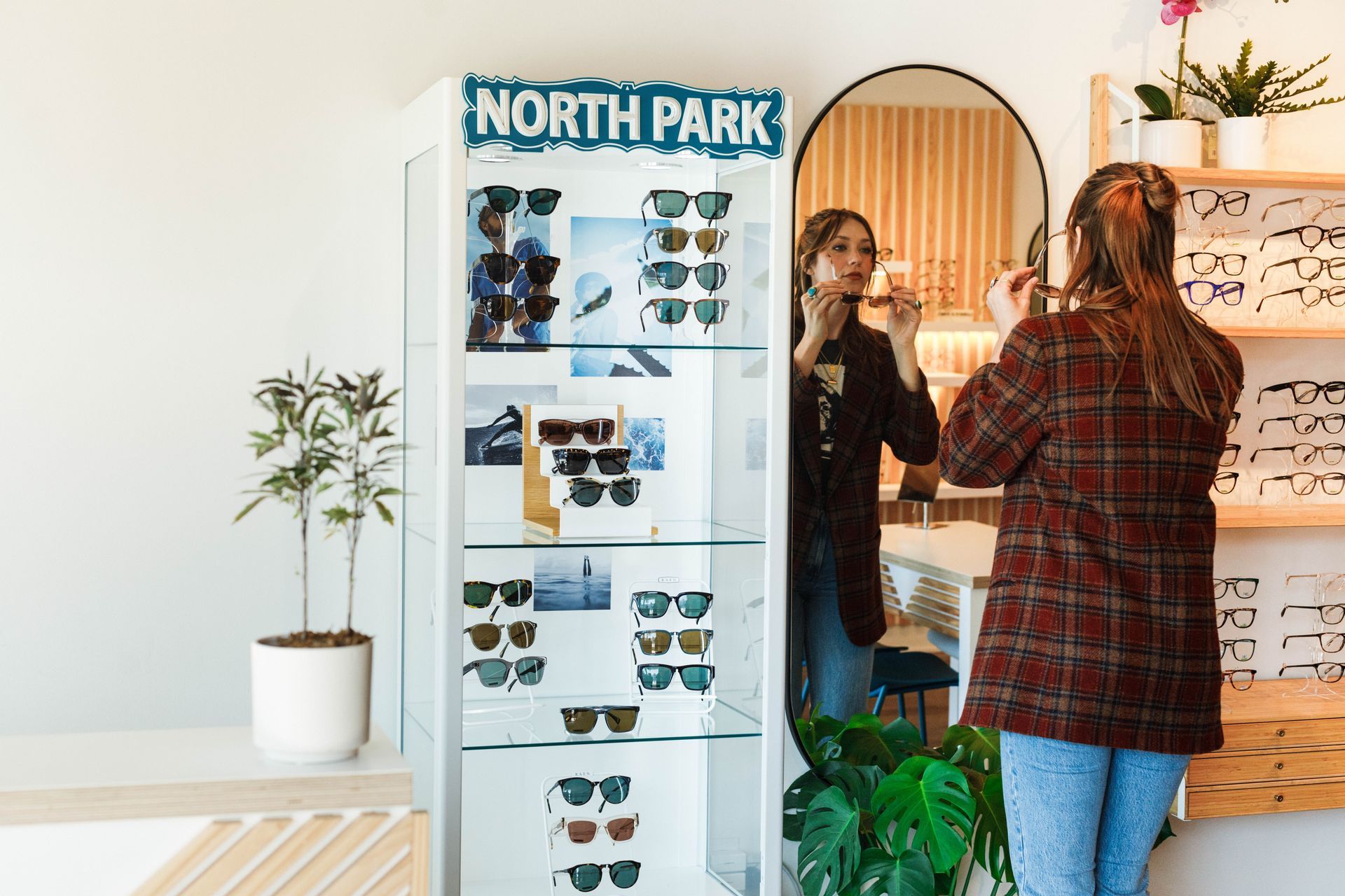 A person in a plaid jacket tries on glasses in front of a mirror next to a retail display case for North Park eyewear.