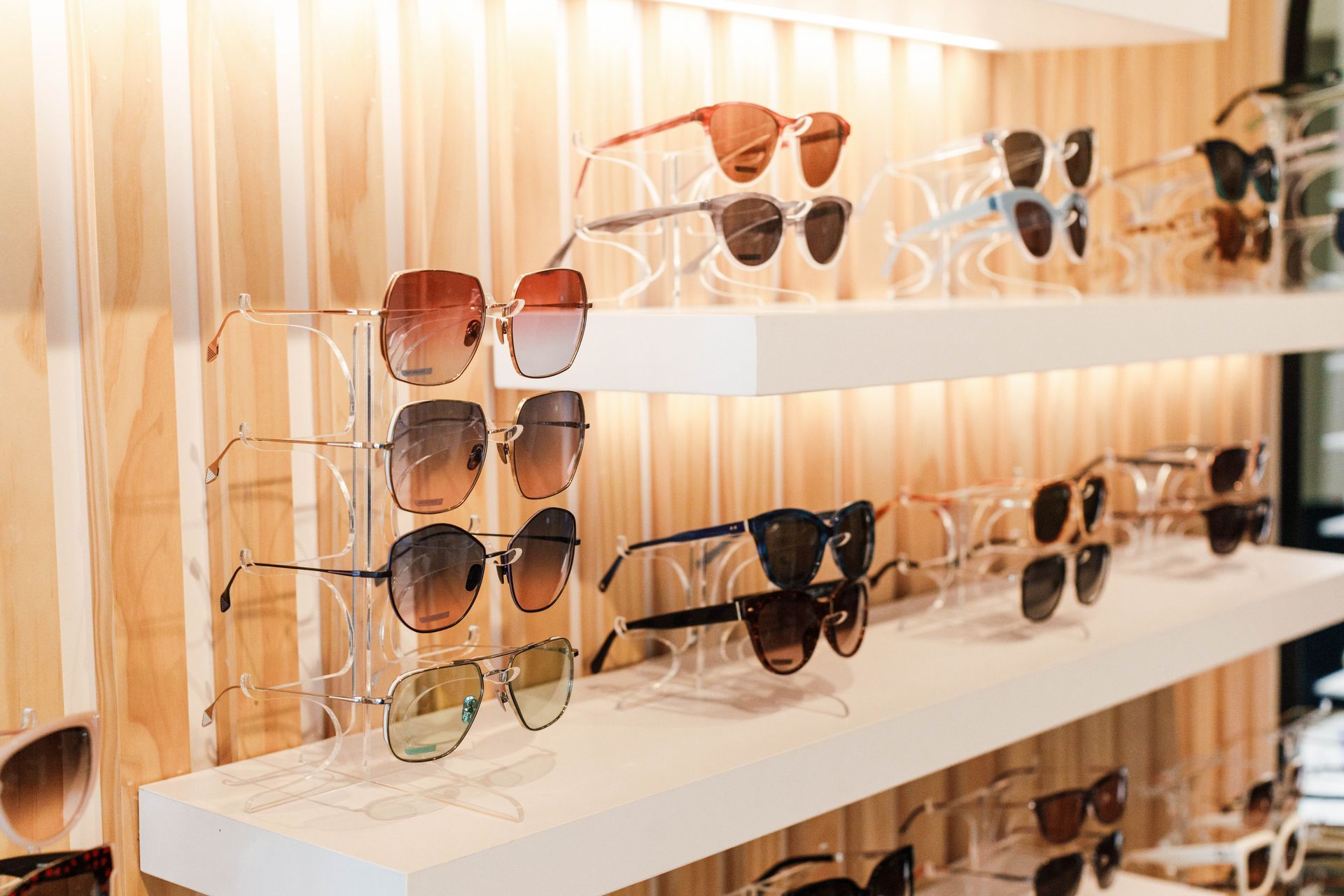 A store display with several shelves holding rows of assorted sunglasses, lit by warm spotlights against wood paneling.