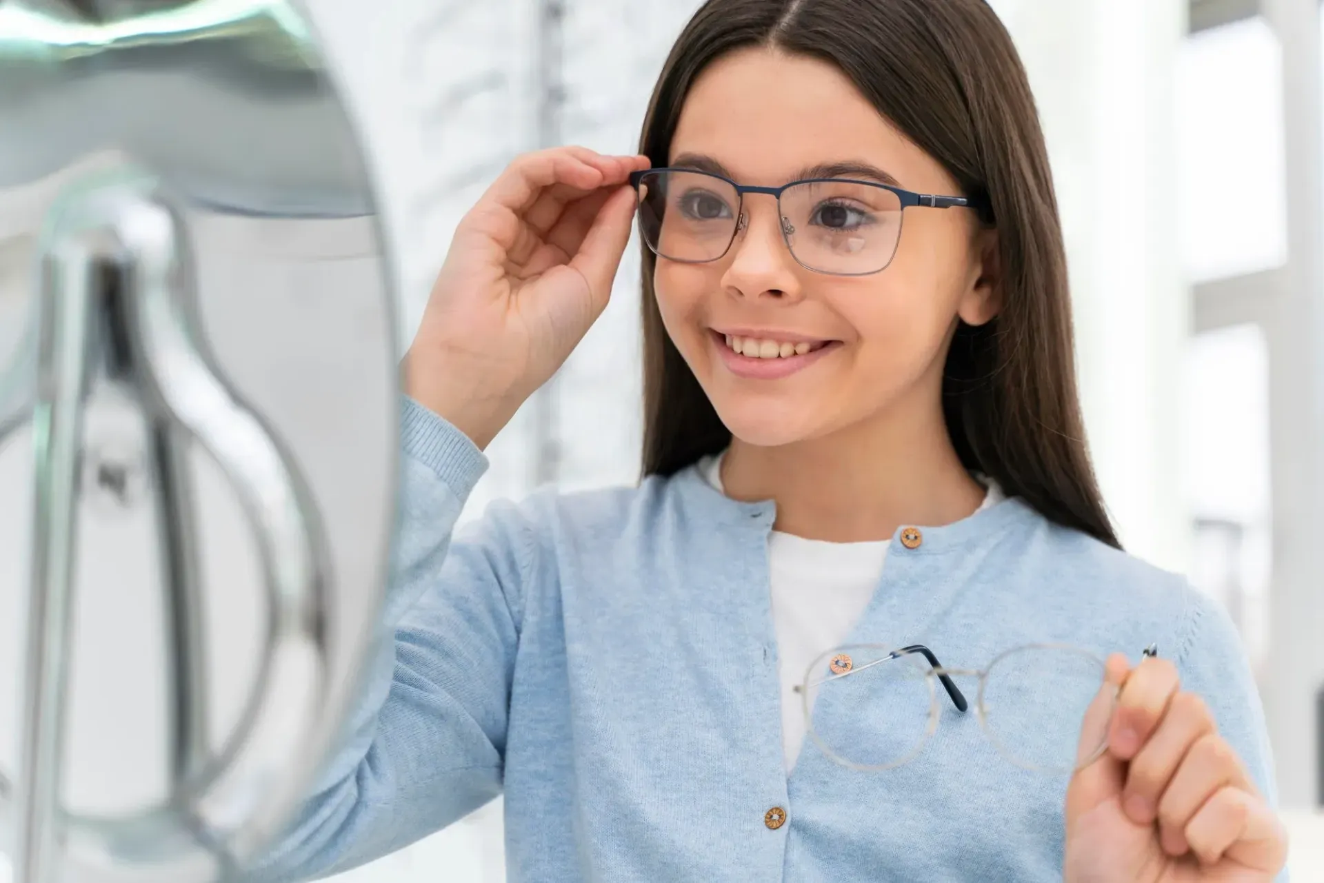 A smiling person tries on new eyeglasses, looking at their reflection in a mirror while holding another pair.