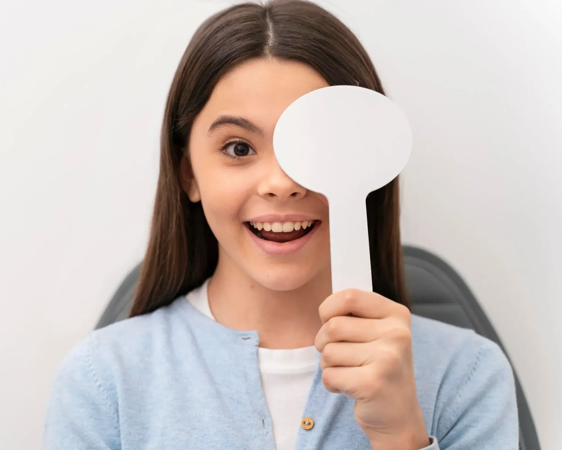 A person smiling while holding a white eye occluder over their left eye during a vision exam against a white background.
