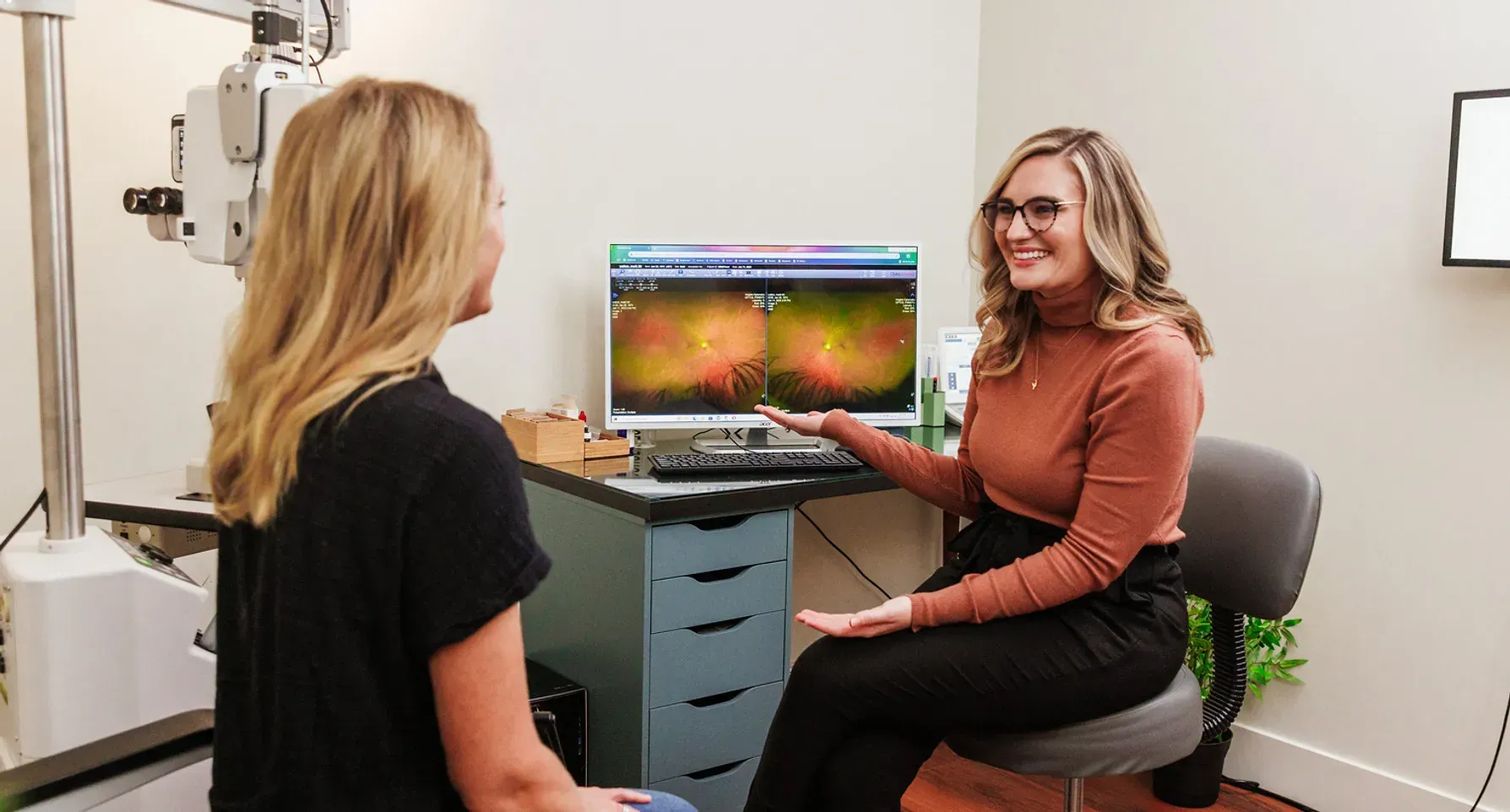 An eye doctor in a rust-colored top reviews retinal scan images on a computer screen with a patient in an exam room.