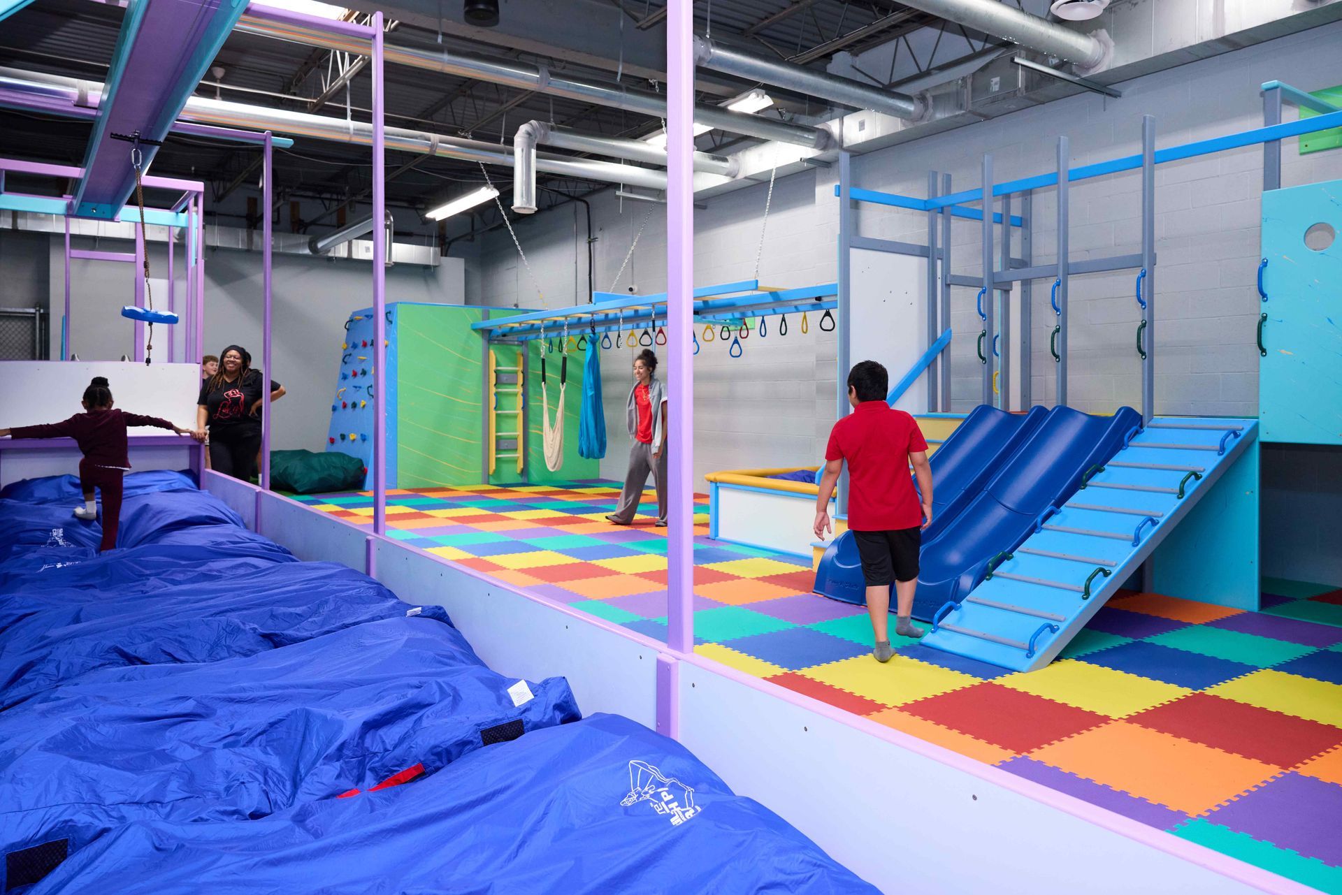 Indoor obstacle course with colorful mats, climbing frames, and people playing on blue and yellow equipment