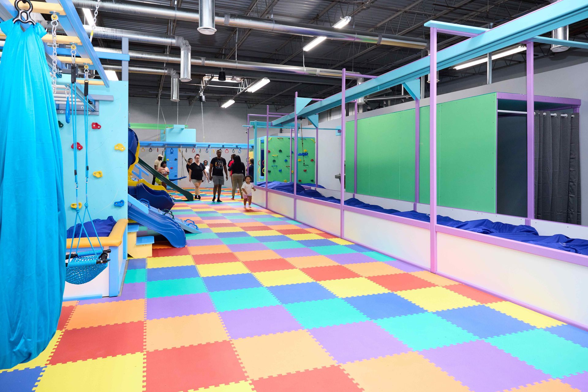 Colorful indoor play area with foam floor tiles, slides, and climbing structures, with people in the background