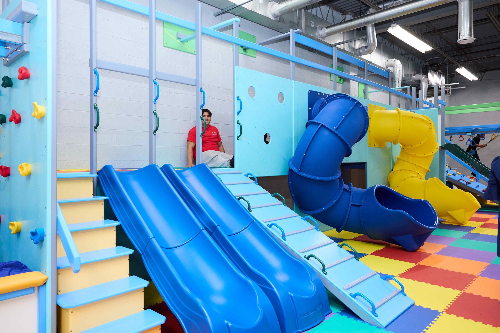 Indoor playground with blue slides, colorful padded floor, and a person standing near the stairs