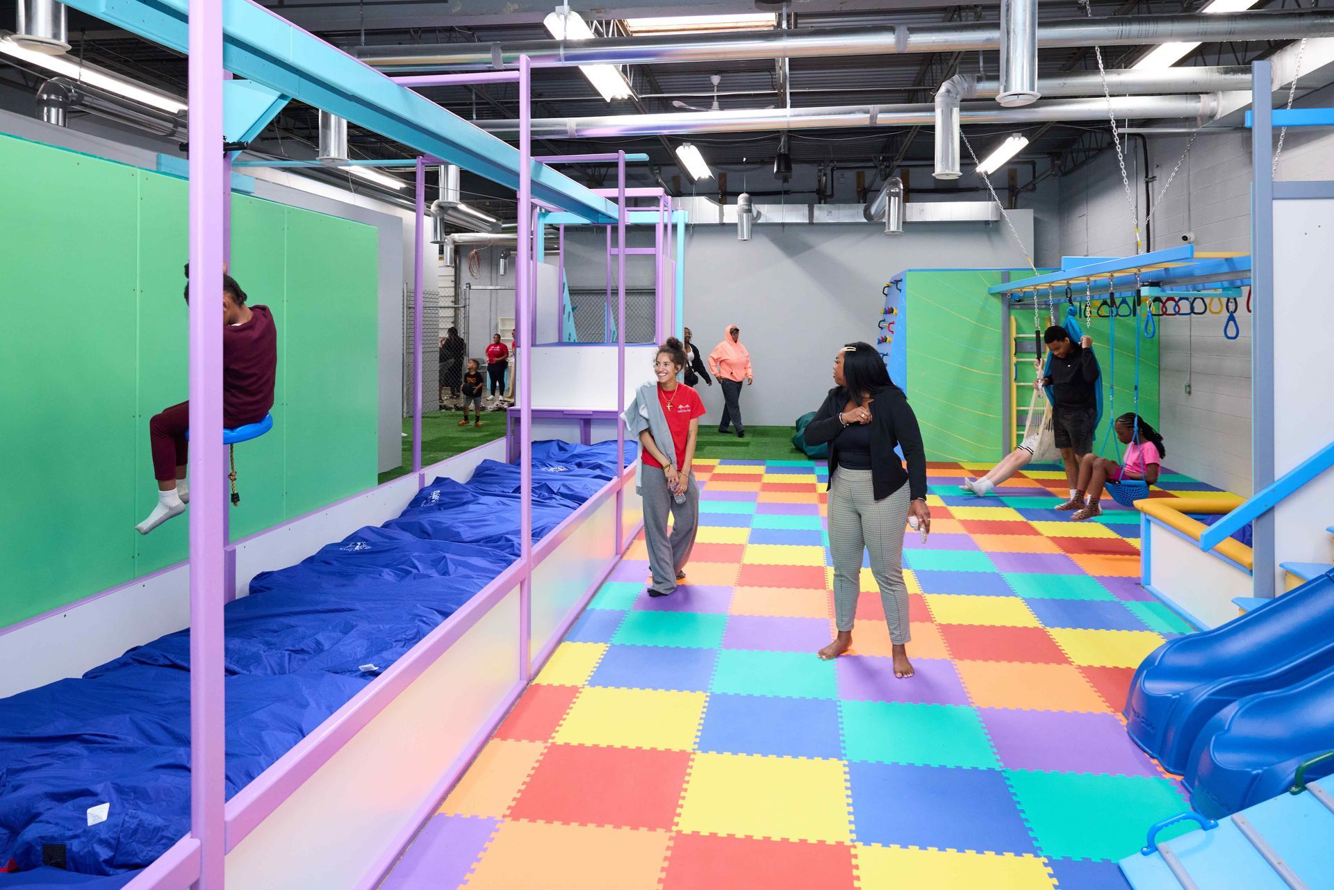 Indoor trampoline play area with colorful foam floor, people bouncing, and blue slide