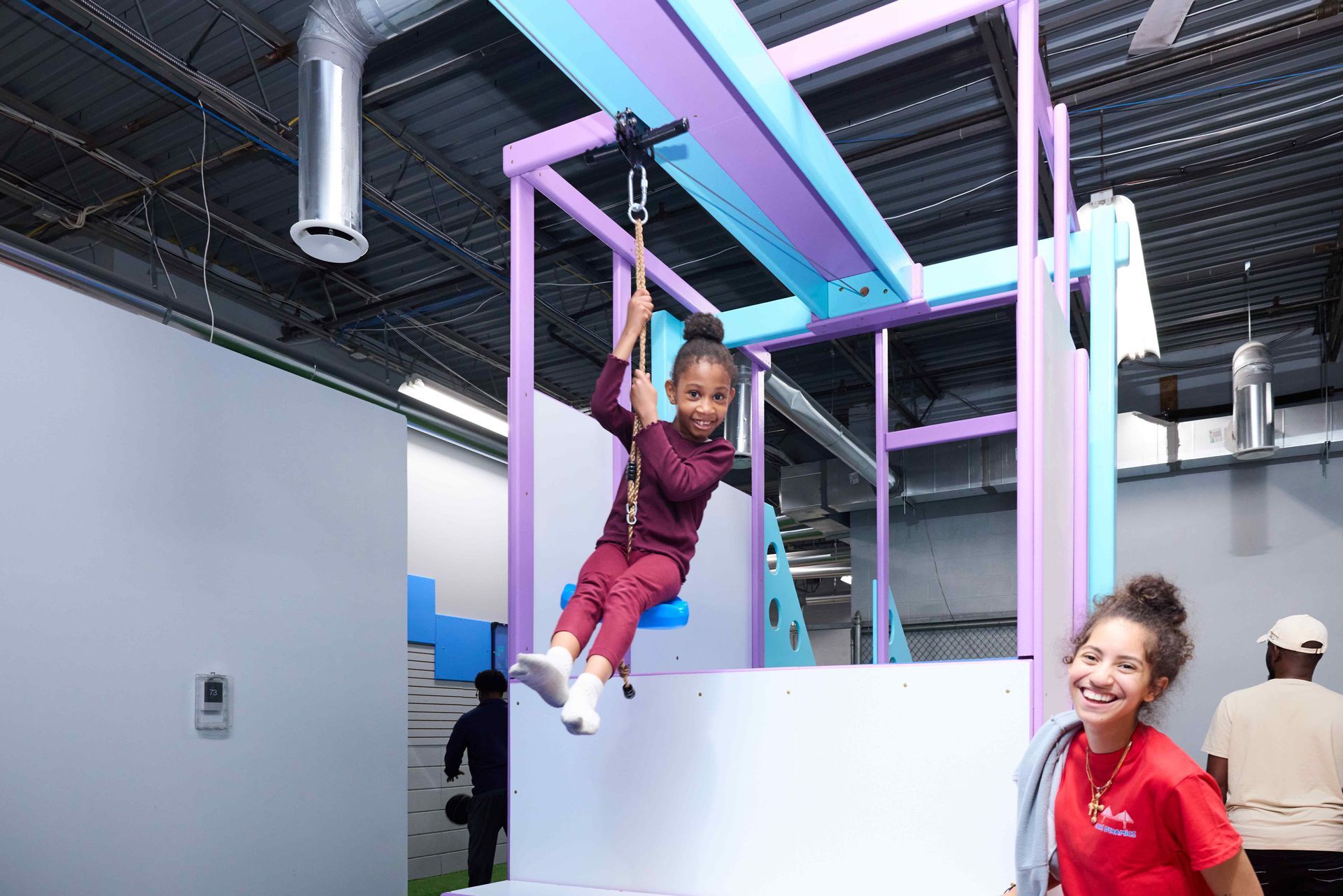Child on colorful indoor playground bars while an adult smiles nearby