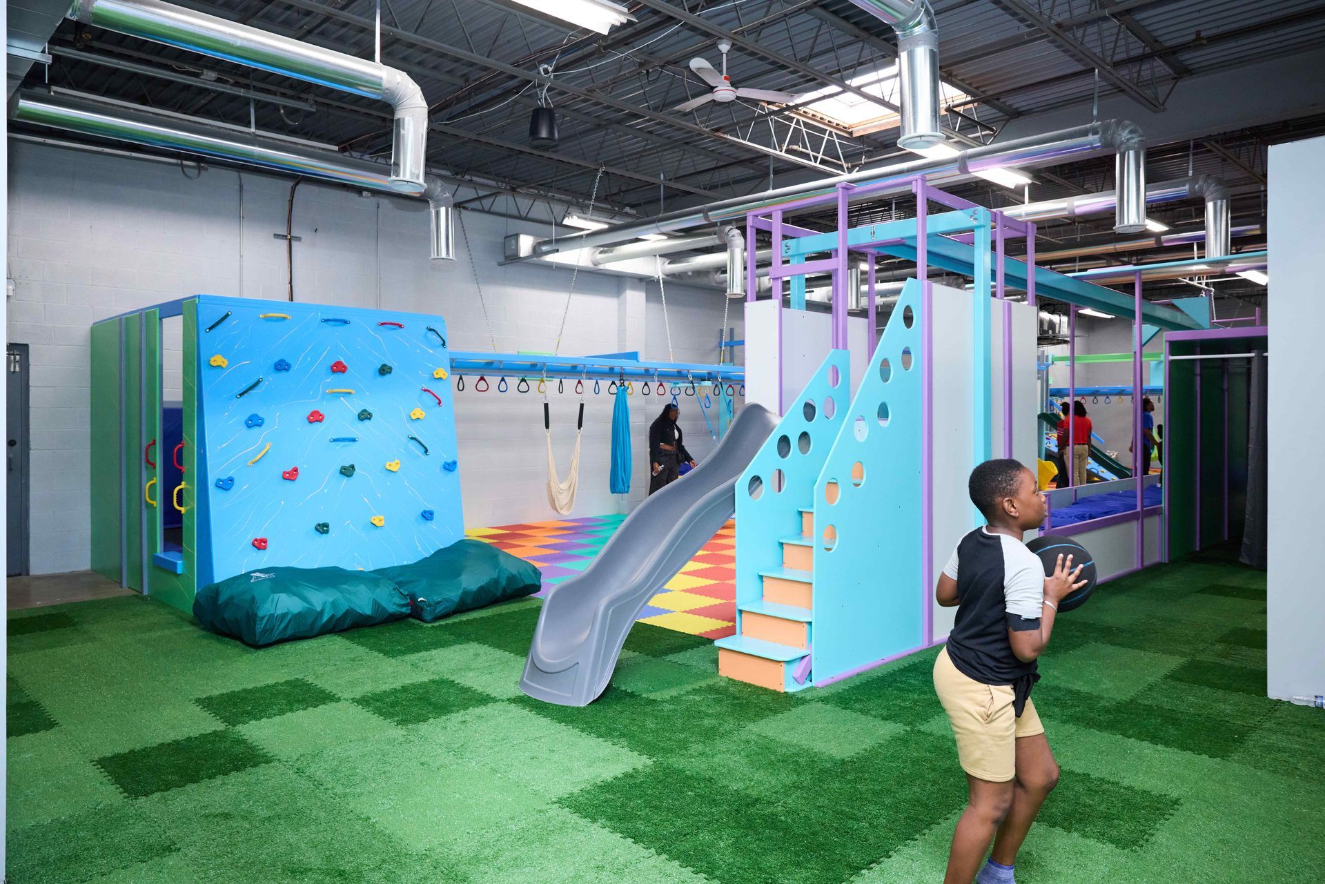Indoor play area with blue climbing wall, slide, and a child standing on green turf