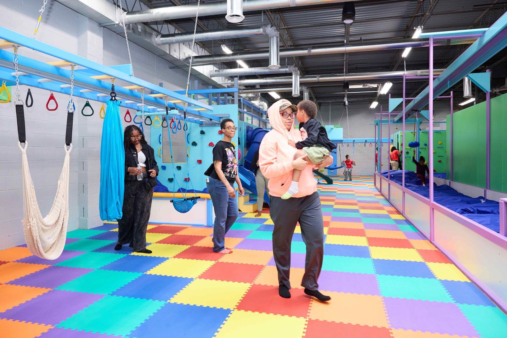 People in a colorful indoor play gym, with a man holding a child on a rainbow foam floor.