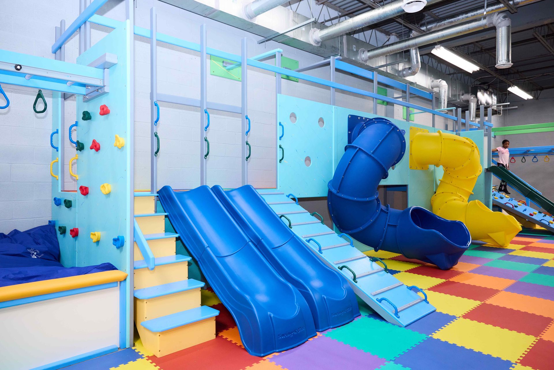 Indoor children's play area with blue slides, climbing wall, and colorful foam flooring