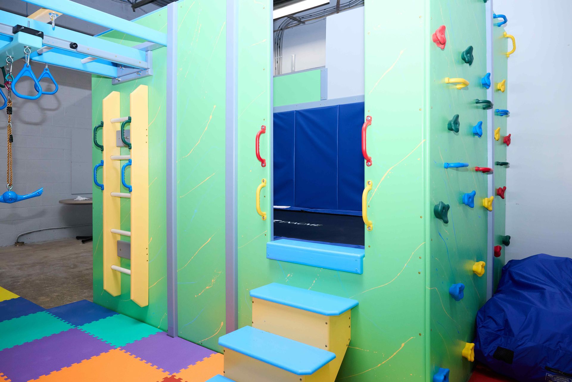 Colorful indoor playroom with climbing wall, steps, padded floor tiles, and hanging rings