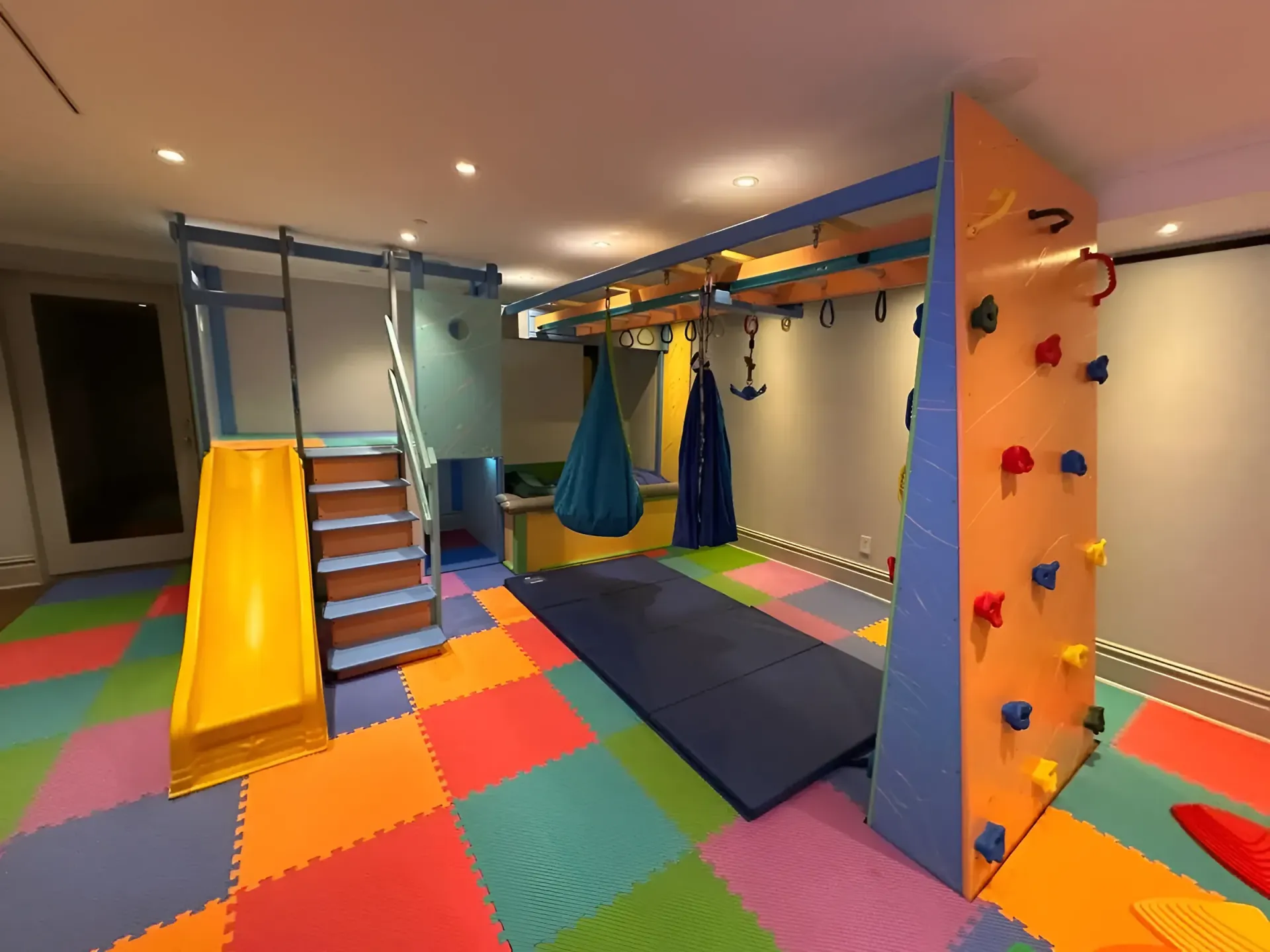 Indoor obstacle gym with colorful foam floor, climbing wall, ropes, slide, and hanging swings