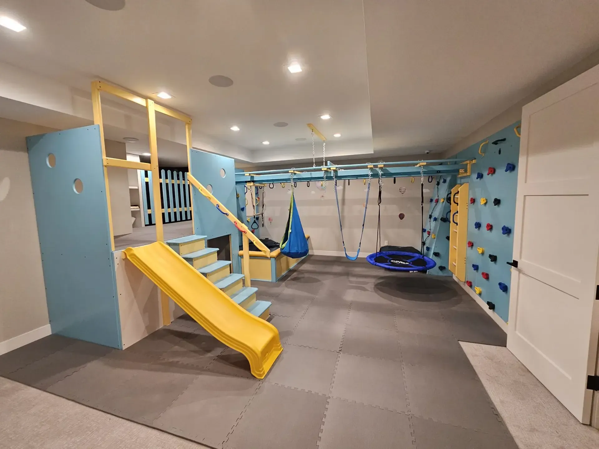 Indoor kids' playroom with yellow slide, climbing wall, rope swing, and padded floor