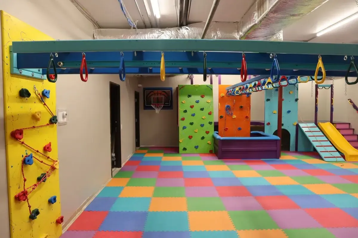 Colorful indoor playroom with foam floor tiles, climbing walls, swings, and a small slide