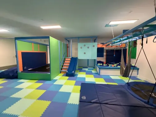 Indoor playroom with colorful foam floor tiles, climbing stairs, hanging rings, and padded equipment