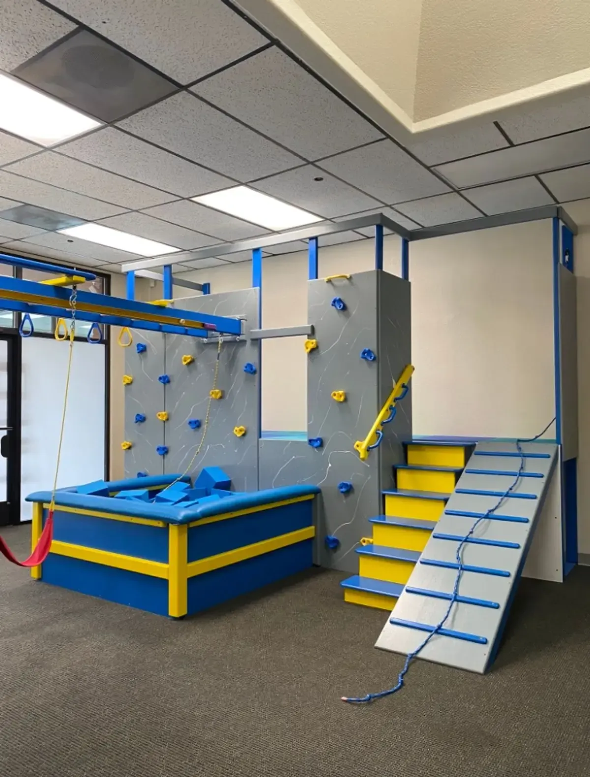 Indoor obstacle course with blue, yellow, and gray climbing features and a ramp in a playroom.