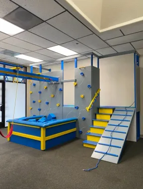 Indoor obstacle course with blue, yellow, and gray climbing features and a ramp in a playroom.
