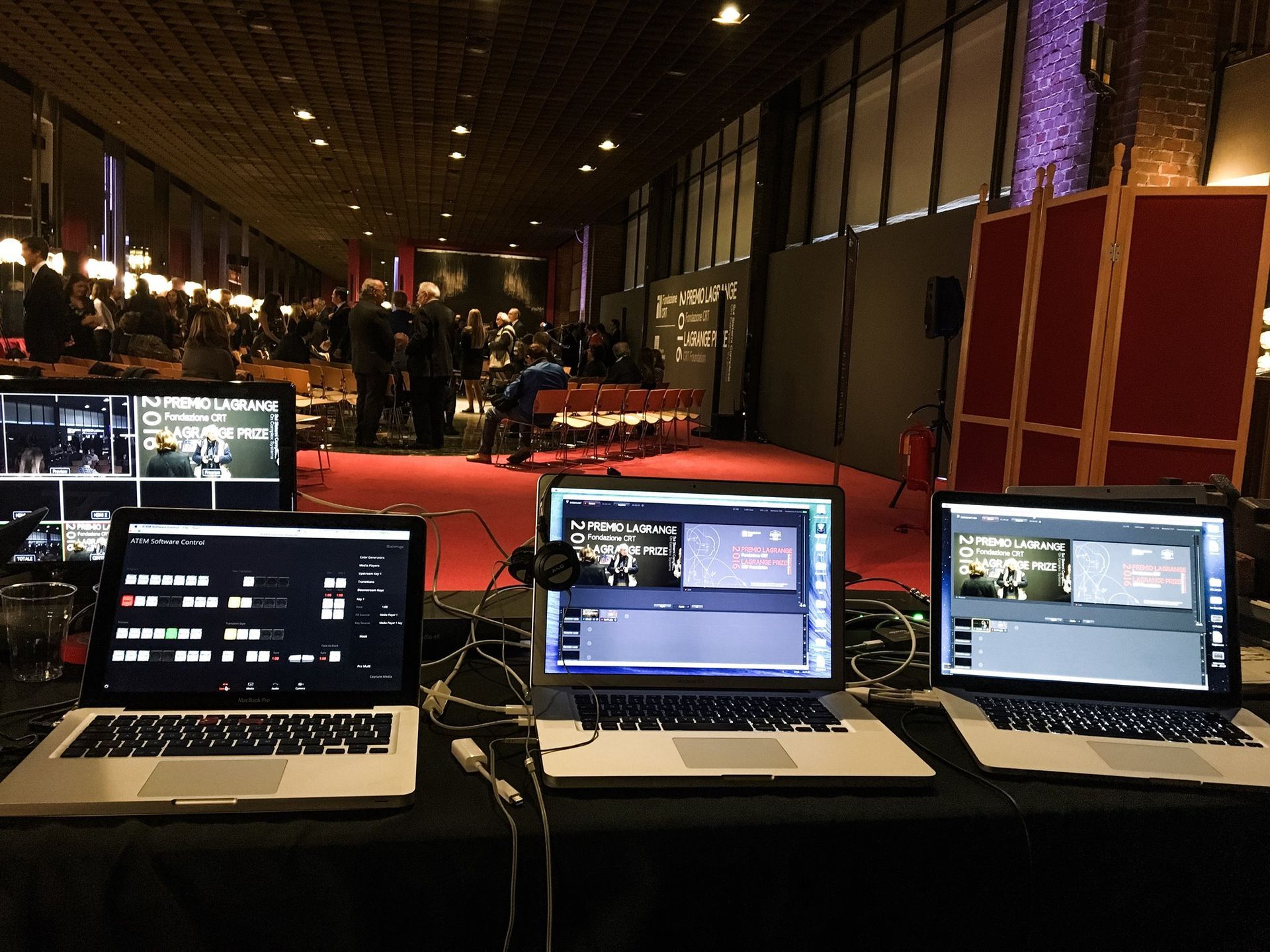 Tre computer portatili su un tavolo con un impianto di streaming live in un locale, con persone sullo sfondo.