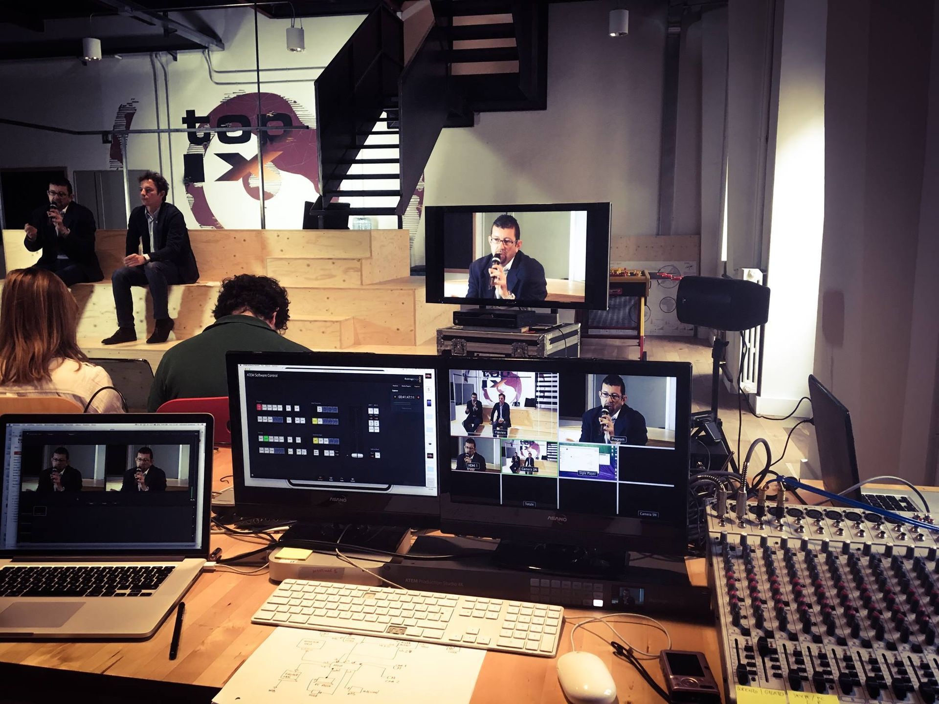 Un evento dal vivo con relatori su un palco, monitor e un mixer audio.
