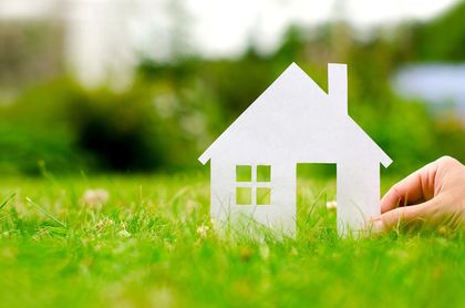 Una mano posiziona un modellino di una piccola casa bianca su un prato verde con uno sfondo naturale sfocato.