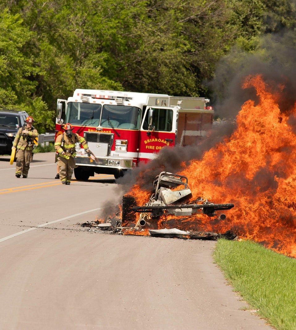 Two fire fighters are walking towards a car fire with a fire hose.
