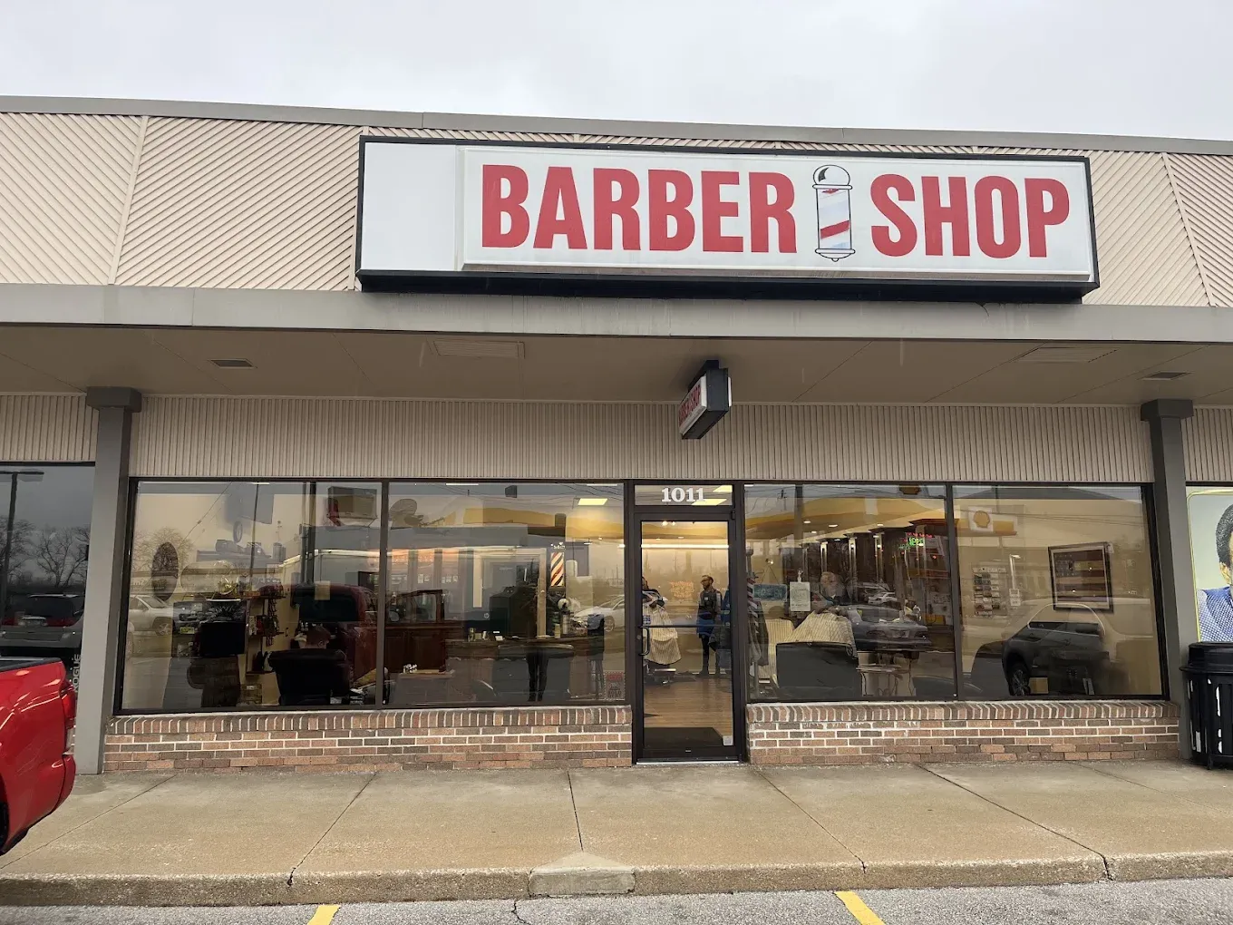A storefront of a barber shop with a red and white sign. Large glass windows show the interior with barber chairs.