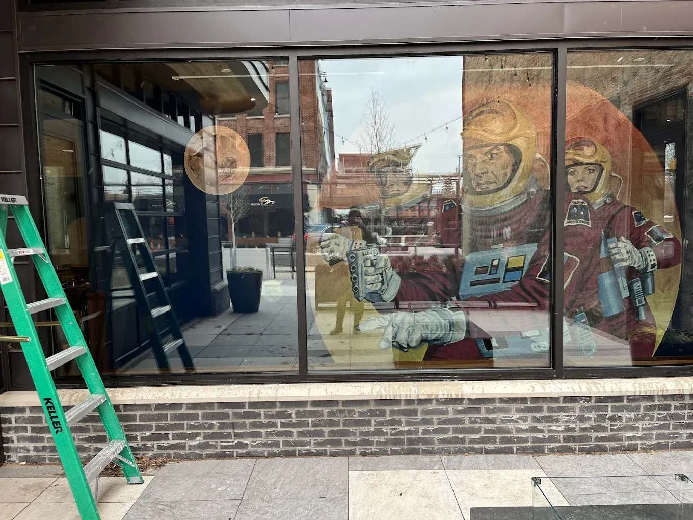 Exterior of a shop with a large window displaying a mural of astronauts. A green ladder leans against the building.