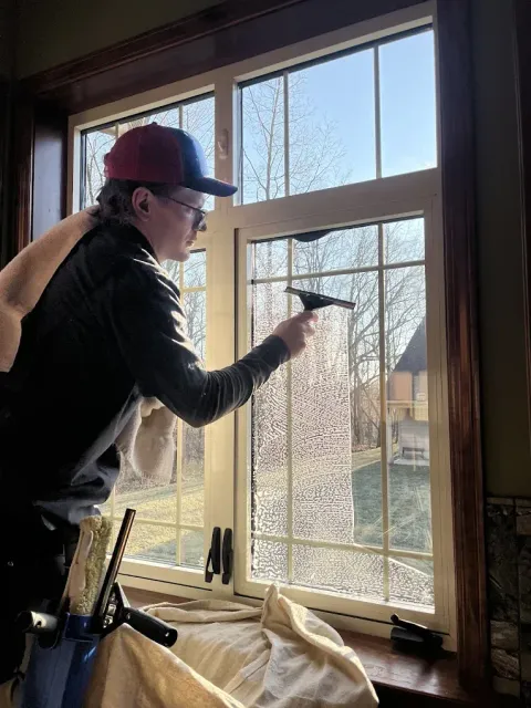 A person cleaning a window with a squeegee. They wear a hat and jacket. The window has a view of a yard and trees.