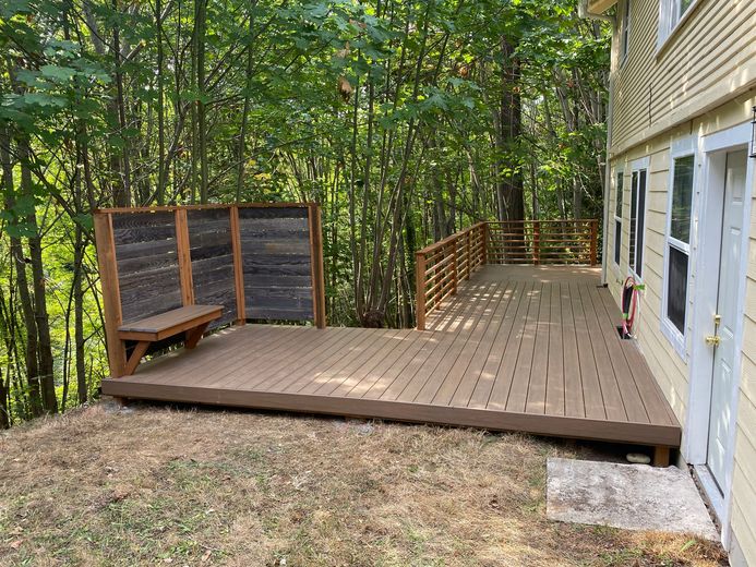 Wooden deck with railings beside a house, overlooking a wooded yard.