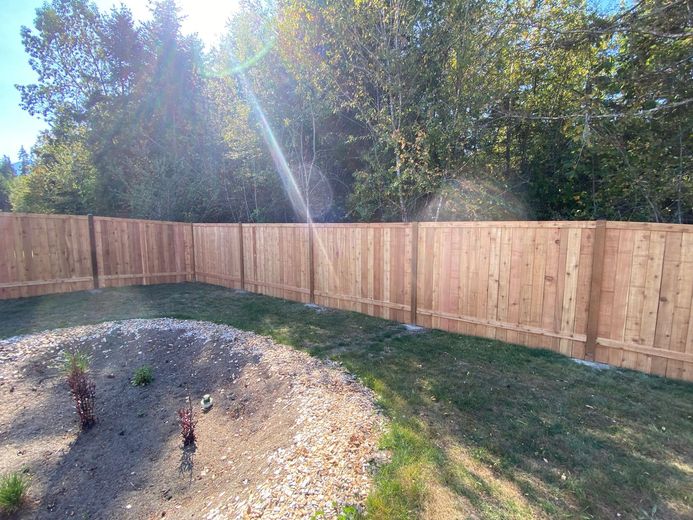 Sunlit backyard with wooden fence, gravel patch, grass, and trees beyond the fence.