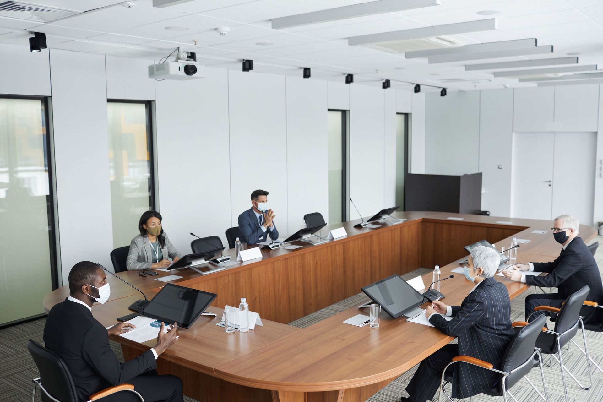 Small conference room meeting with several people seated around a curved wooden table with laptops
