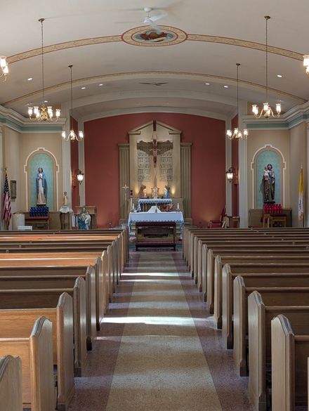Church interior with wooden pews, central aisle, and altar beneath ornate arches and chandeliers