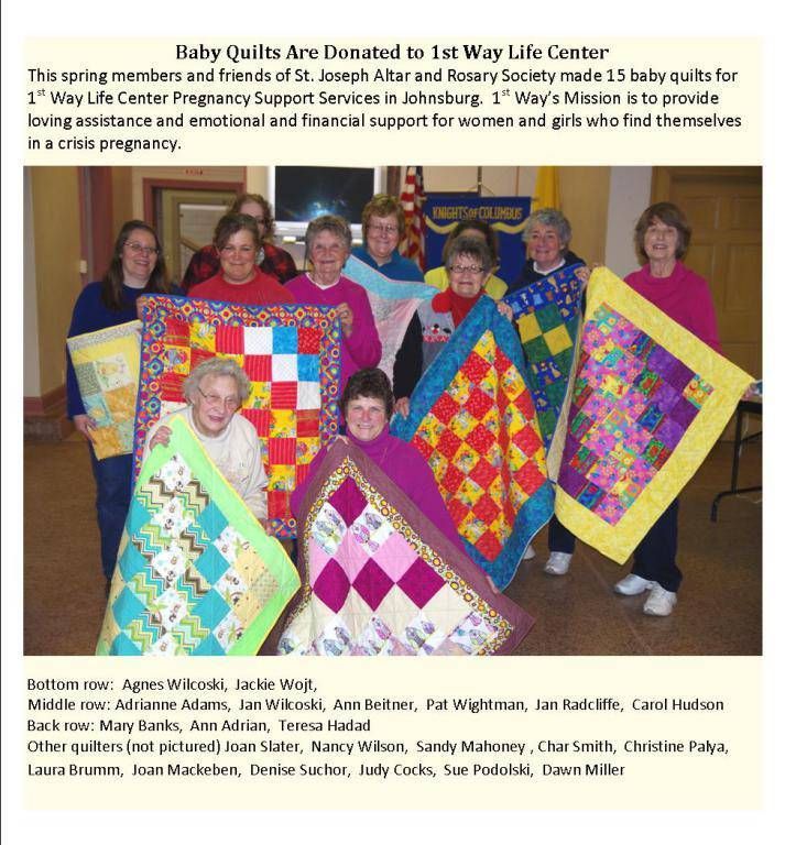Group holding colorful patchwork quilts in a community room, smiling for a fundraiser photo