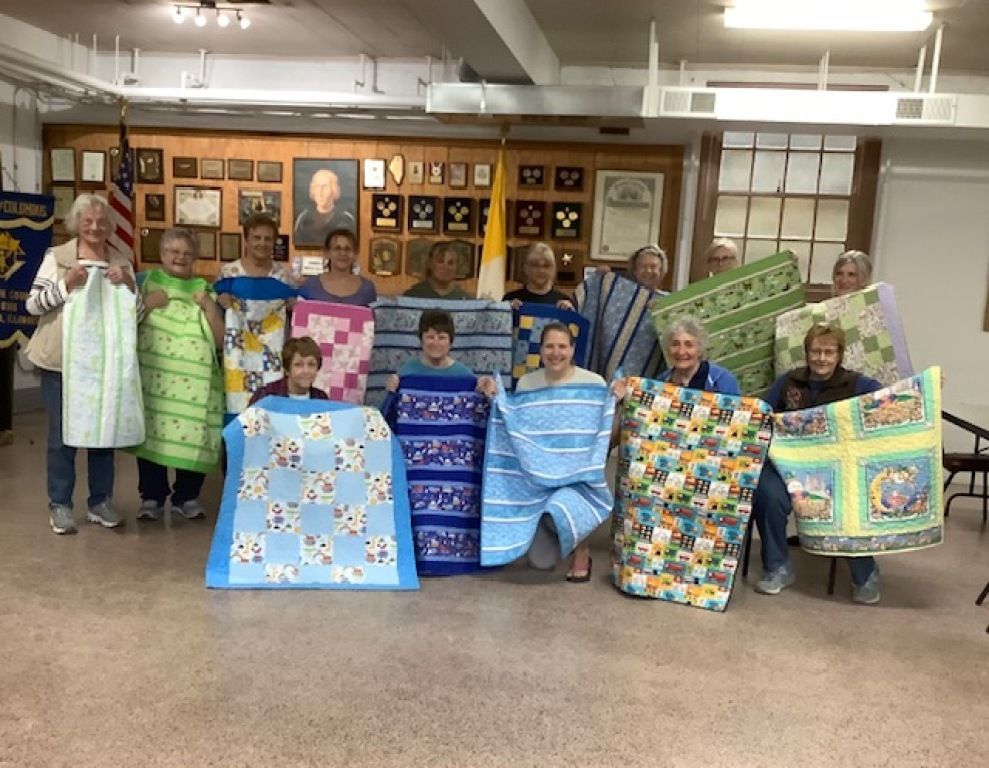 Group of people holding colorful quilts in a community room with plaque wall display.