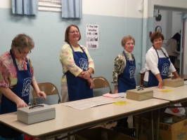 Four women in aprons stand behind a long table with bowls and supplies in a bright room