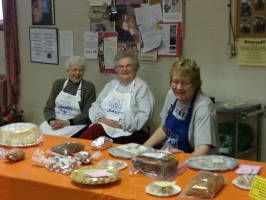 Three women sit behind a table of food at a community event, smiling at the camera.