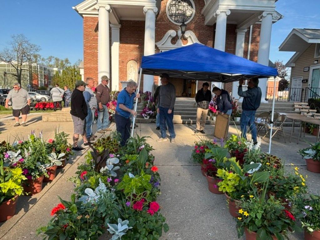 Outdoor flower sale with colorful potted plants and people under a blue canopy in front of a brick building