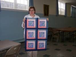 Person holding a large quilt with blue and pink square patterns in a room with tables and chairs.
