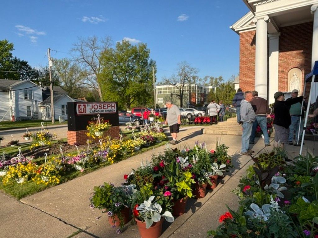 People browsing colorful flower baskets and plants outside a brick building on a sunny day